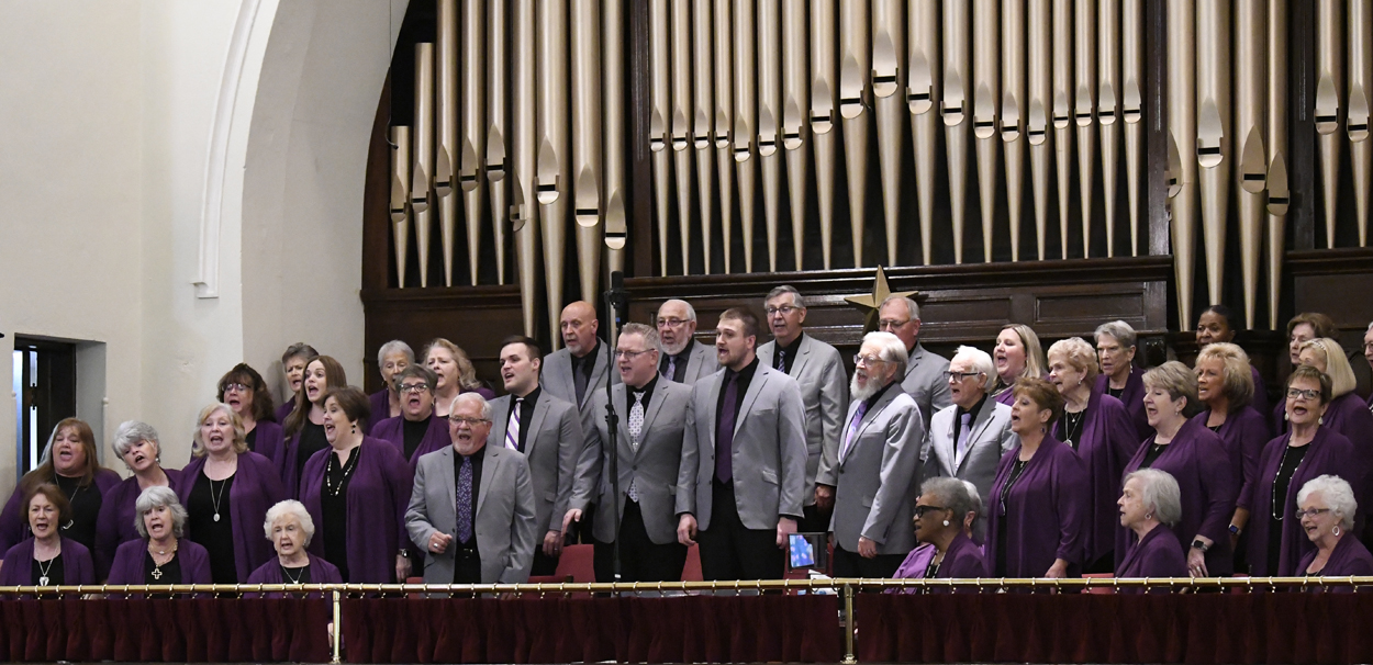 A choir dressed in gray and purple sings in front of a large church organ during a performance.