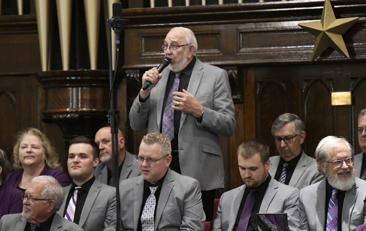 older man in a gray suit sings into a microphone while standing among seated men in matching suits inside a formal setting.