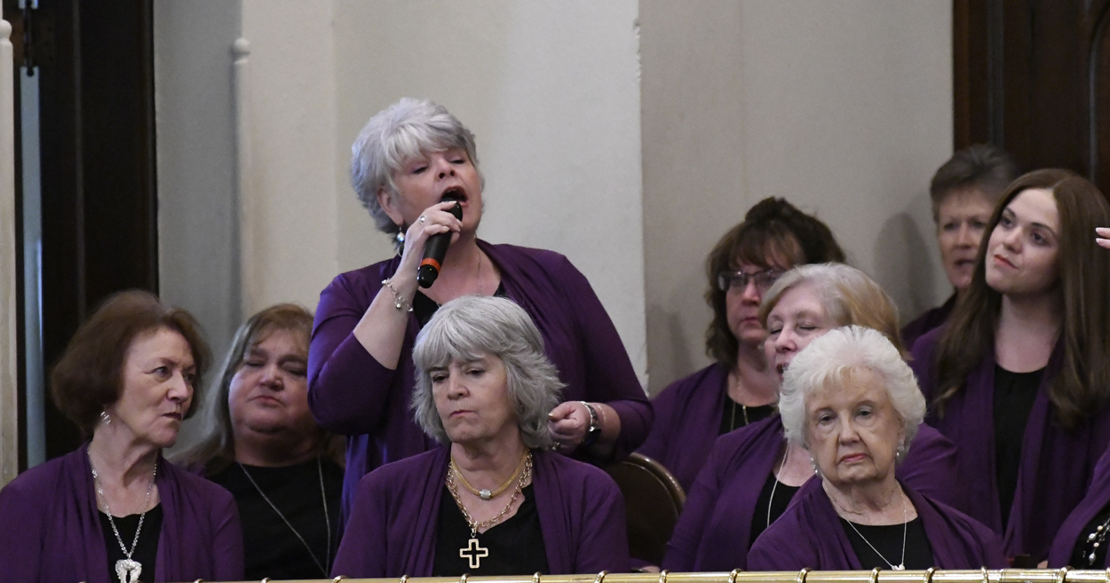 A group of women, mostly with gray hair, wear purple cardigans; one woman stands singing into a microphone, while others sit and listen attentively.