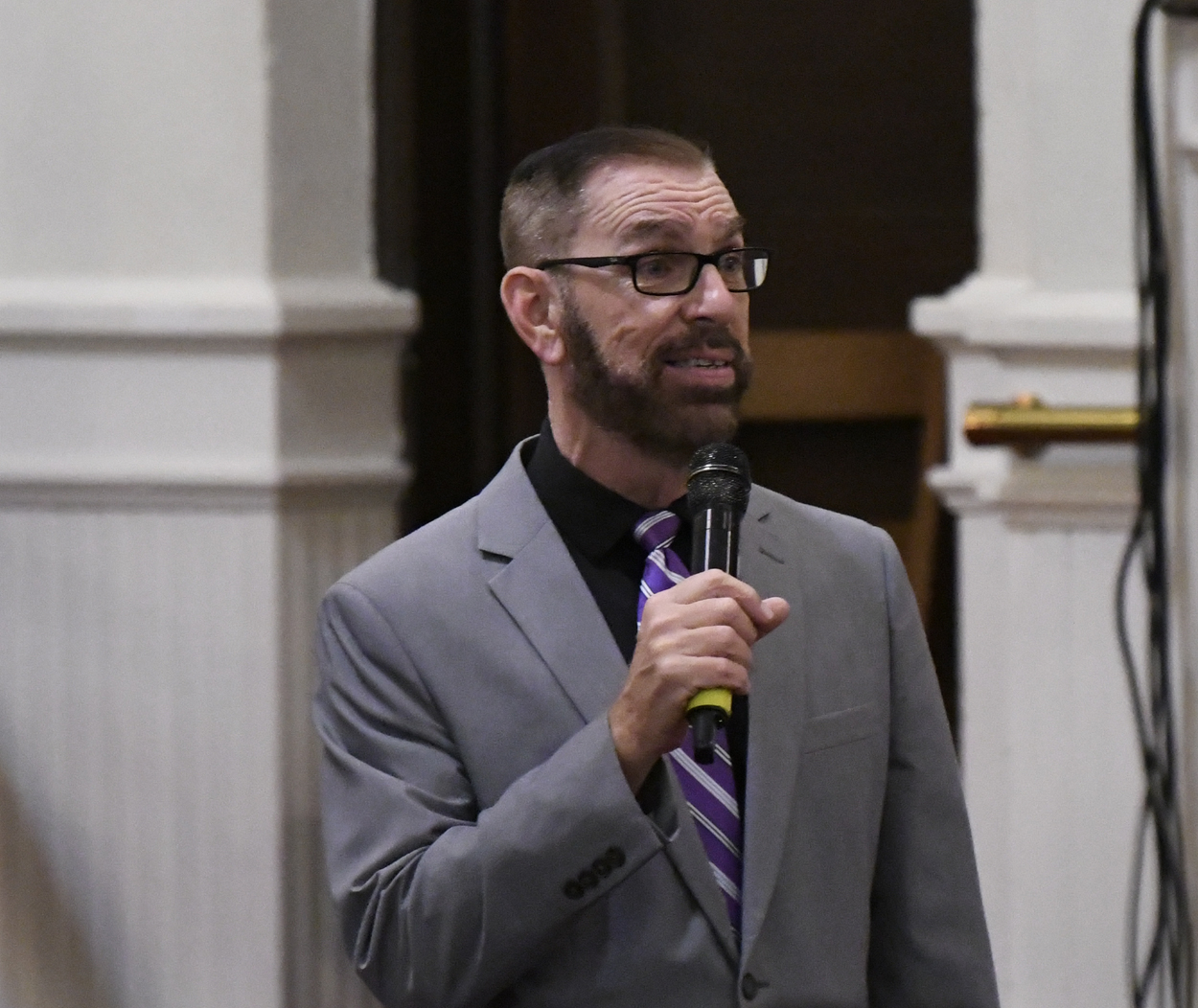 Man with glasses and beard wearing a gray suit holds a microphone while speaking indoors.