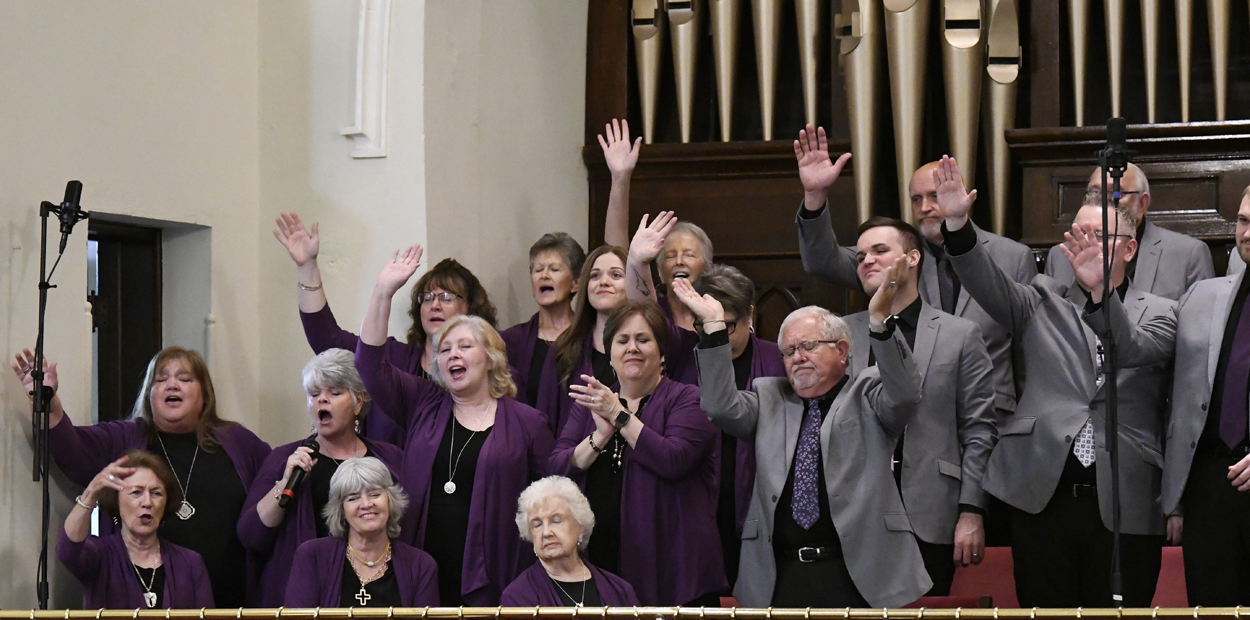 A choir of older adults sings joyfully, many raising their hands, dressed in purple attire, standing in front of organ pipes in a church setting.