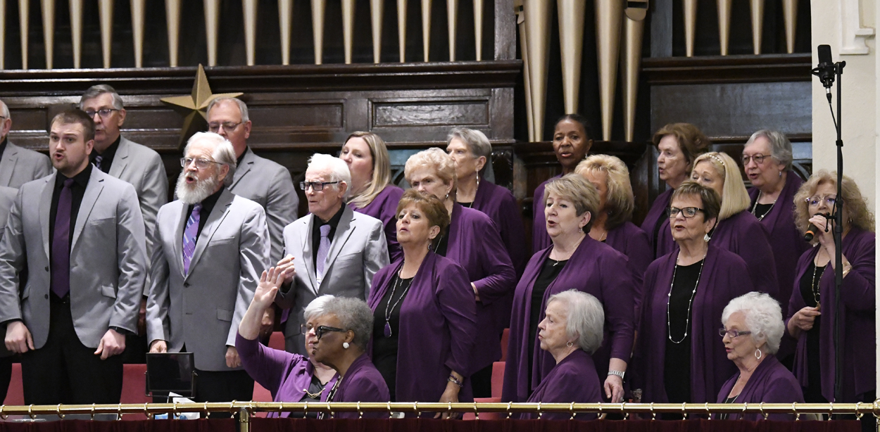 A choir of men and women, mostly wearing purple jackets, sings in front of an organ in a church setting.