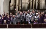 A choir of men in gray jackets sings enthusiastically on stage in front of large organ pipes.