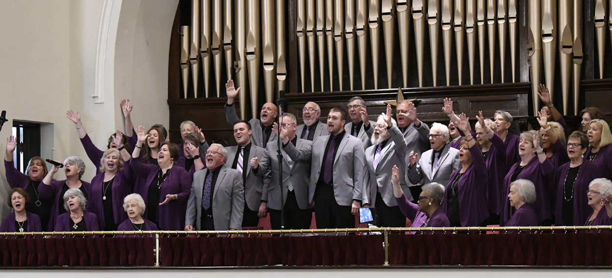 A choir of men in gray jackets sings enthusiastically on stage in front of large organ pipes.