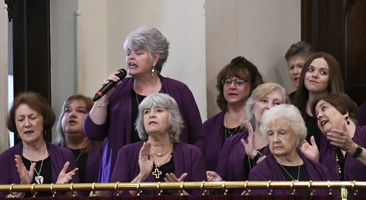 A group of women in purple outfits sing together, one holding a microphone while others stand and clap in what appears to be a choir setting.