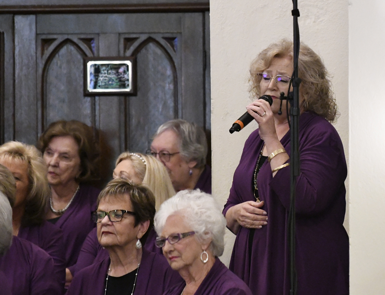 A group of older women in matching purple outfits, one standing and singing into a microphone, others seated, indoors near a wooden door.