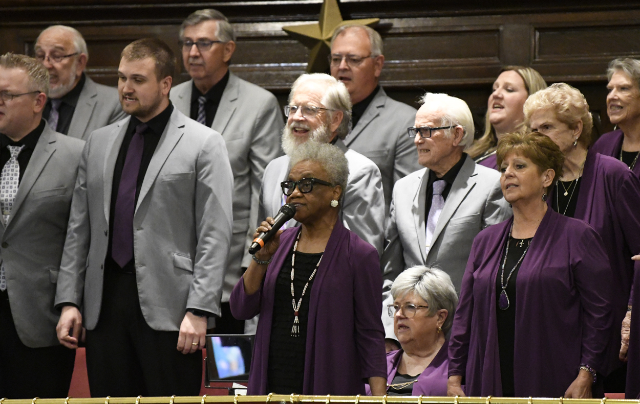 A diverse group of choir singers dressed in gray suits and purple attire perform on stage, with one woman in front holding a microphone.