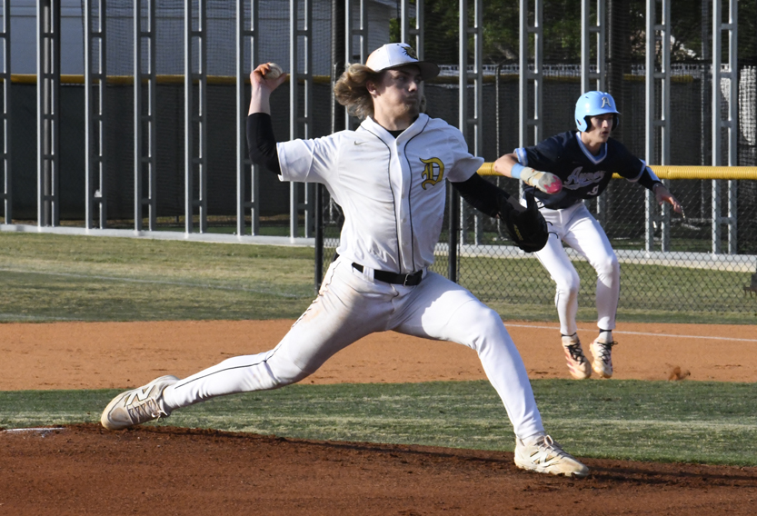 Baseball pitcher in white uniform mid-throw on the mound, while a base runner in blue prepares to run. Playing on a sunny outdoor field.