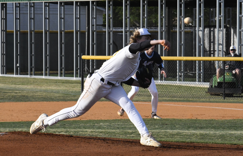 A baseball pitcher in a white uniform throws a pitch on a sunny field, with another player and a person sitting in the background.