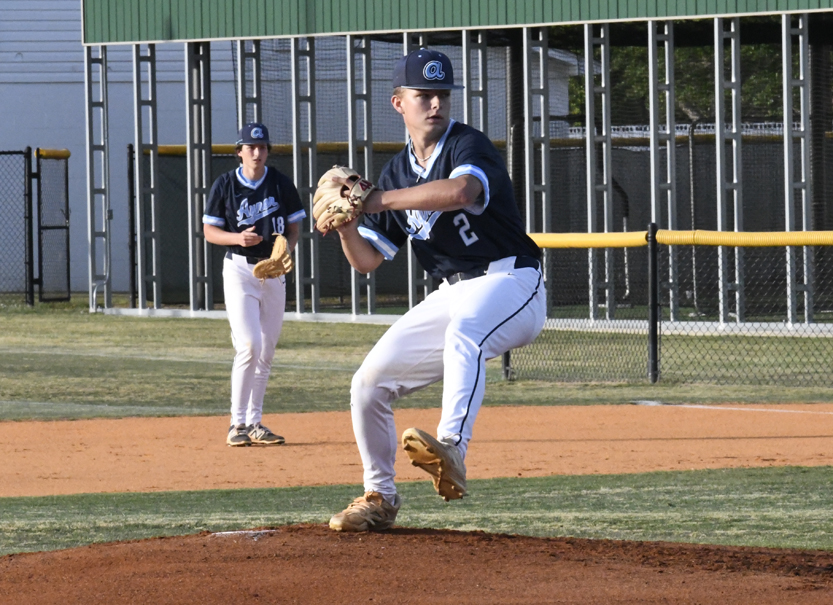A baseball pitcher in blue and white uniform prepares to throw the ball on the mound, with a teammate standing in the background on the field.
