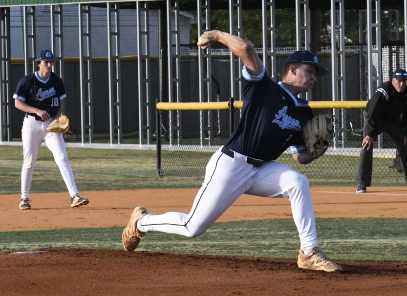 A baseball pitcher in a navy uniform throws a pitch on a field, with a teammate and umpire visible in the background.