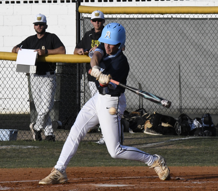 Baseball player in blue helmet swings bat, hitting the ball during a game, with two coaches and equipment visible in the background.