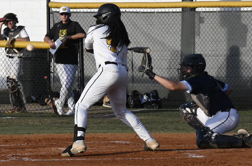 A baseball player in white uniform swings at a pitched ball while the catcher and teammates watch from behind the fence.