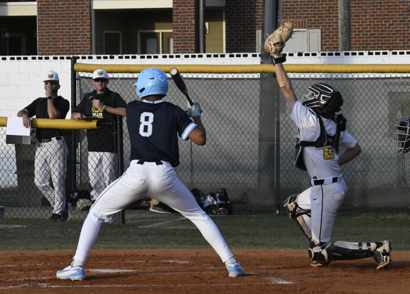 A baseball batter prepares to swing as the catcher raises his glove, with coaches and players watching from behind the fence.