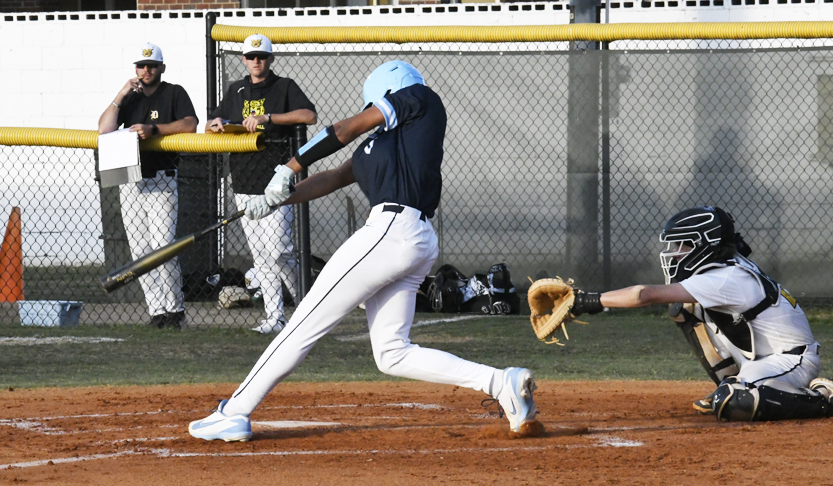 A baseball player in blue swings at a pitch while the catcher in white and black squats behind home plate, with coaches and spectators in the background.