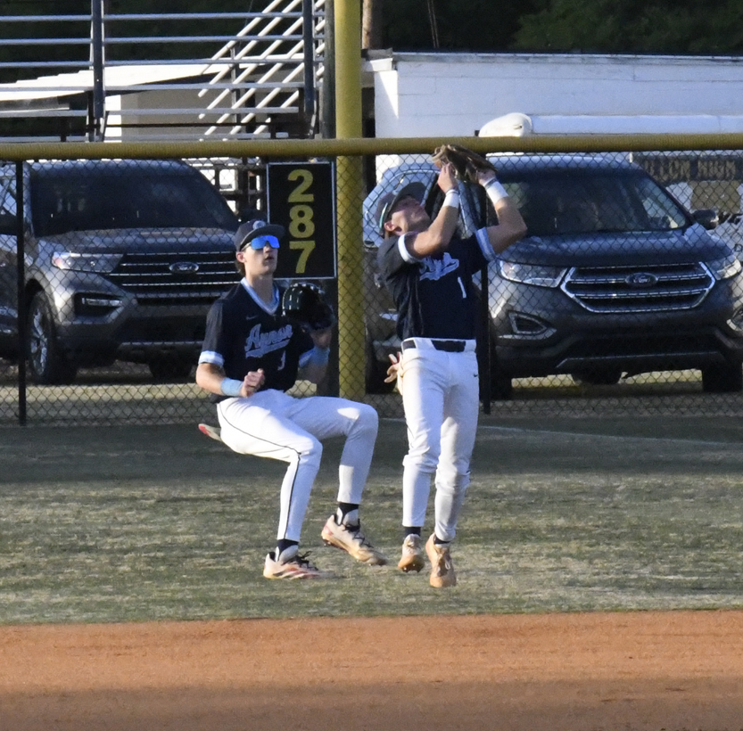 Two baseball players in navy uniforms converge in the outfield as one prepares to catch a fly ball near the fence during a game.