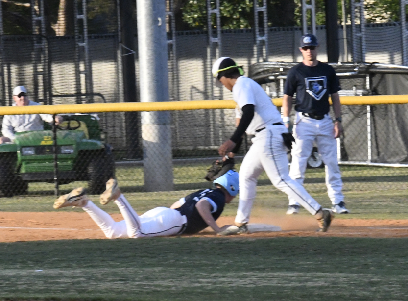 Baseball player slides to base as opposing player attempts to tag, with spectators and another player watching in the background.