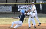 A baseball player in blue slides into base as a player in white tries to tag him, with another player watching in the background.