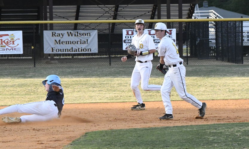 A baseball player in a blue helmet slides into base as two players in white uniforms prepare to catch and throw the ball.