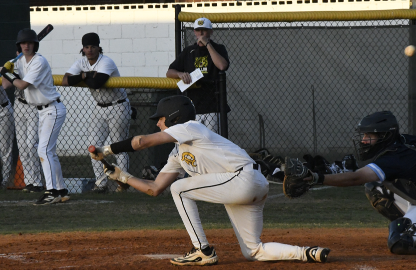 A baseball player bunts the ball during a game, while the catcher and teammates watch from behind the fence.