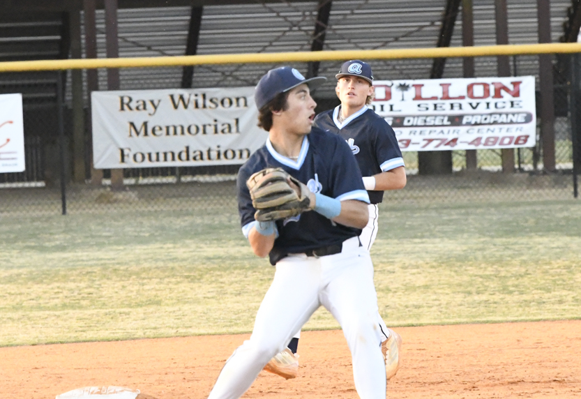 Two baseball players in blue uniforms on a field, one preparing to throw a ball while the other watches in the background.