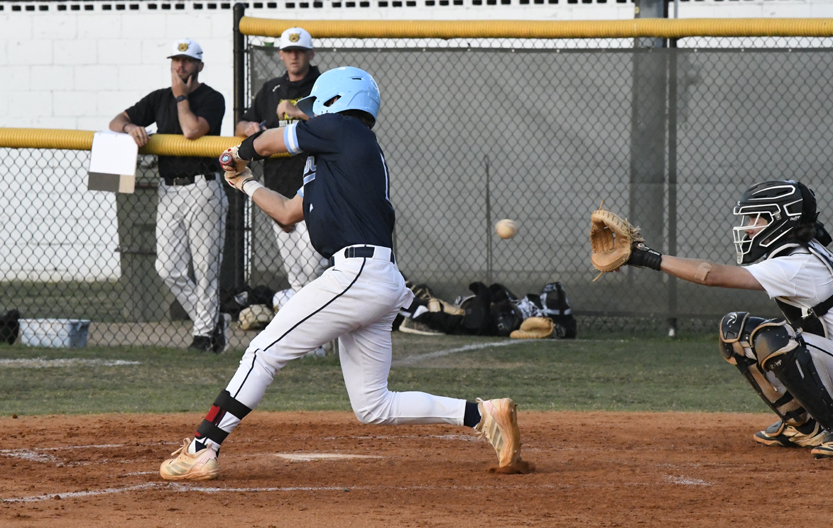 Baseball player in blue helmet swings at a pitch, catcher reaches out to catch the ball, two coaches watch from behind the fence.