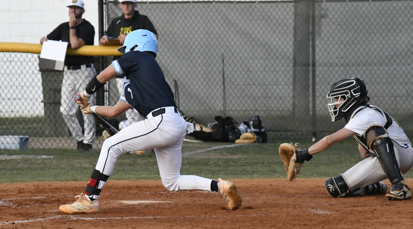 Baseball batter in blue swings at a pitch while catcher in white gear reaches to catch the ball; two people watch from behind the fence.