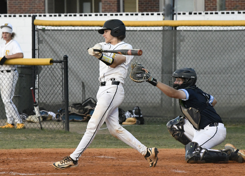 A baseball player in white uniform batting, with a catcher in blue gear ready behind home plate, during a game on an outdoor field.