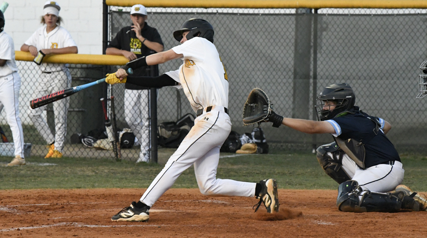 A baseball player in white swings at a pitch while the catcher in dark gear prepares to catch. Spectators and teammates watch from behind a fence.