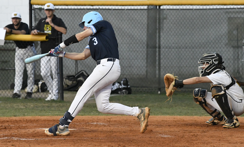 Baseball player in blue uniform swings bat at pitch, while catcher in white gear catches behind home plate; players and fence visible in background.