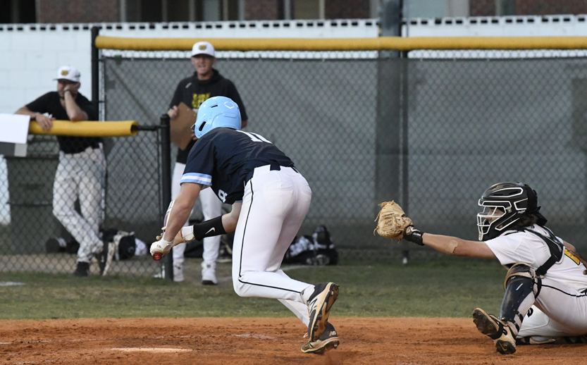 A baseball player in blue bunts the ball while a catcher in white reaches out with a glove. Players and fence are visible in the background.