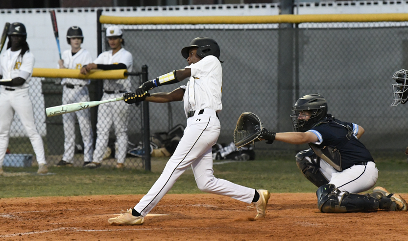 A baseball player in white swings at a pitch as the catcher and teammates watch during a game on a dirt field.