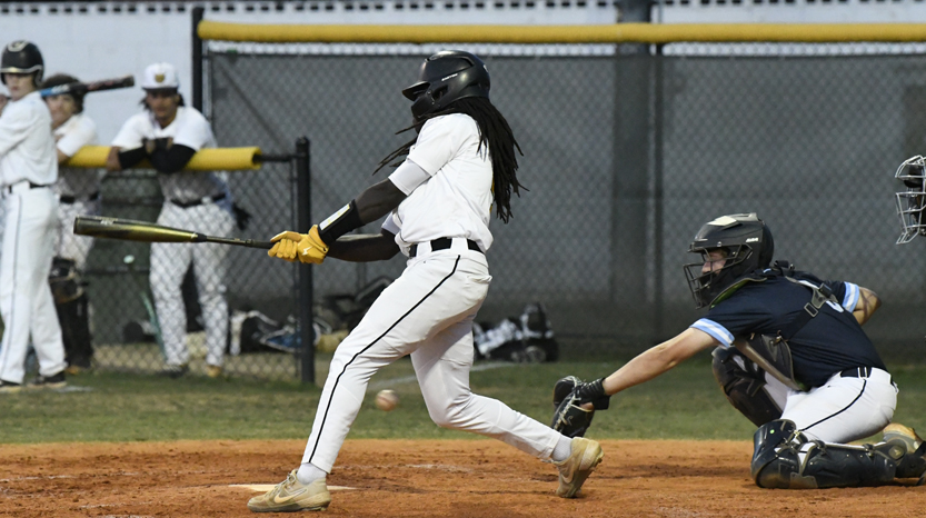 Baseball player in white uniform swings bat at pitch, catcher in dark gear crouches behind home plate, teammates watch from dugout in background.