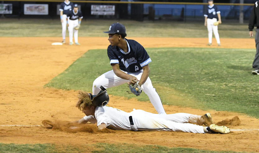 Baseball player slides into base as opposing player prepares to tag, infield dirt flying, other players and umpire visible in the background.