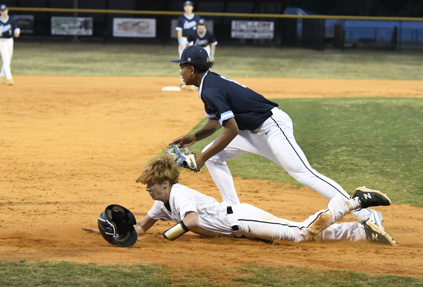 A baseball player in white slides into base as an opposing player in navy attempts to tag him out during a game on a dirt field.