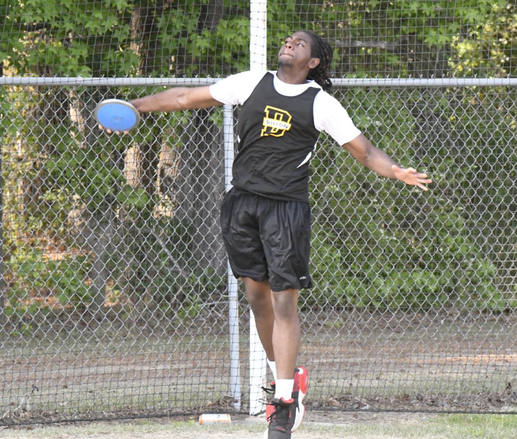 A male athlete in sportswear prepares to throw a discus at an outdoor track and field event, with a chain-link fence and trees in the background.