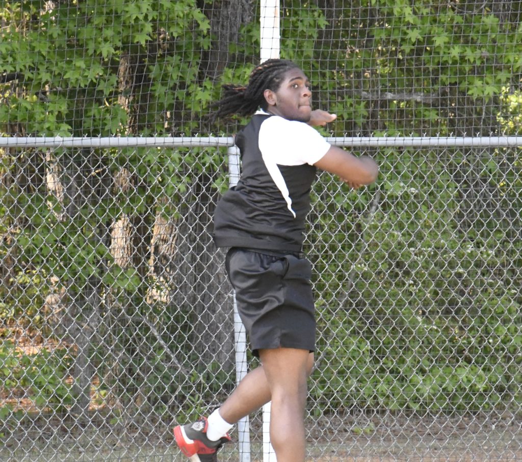 Athlete in sportswear prepares to throw a discus behind a chain-link fence with trees in the background.