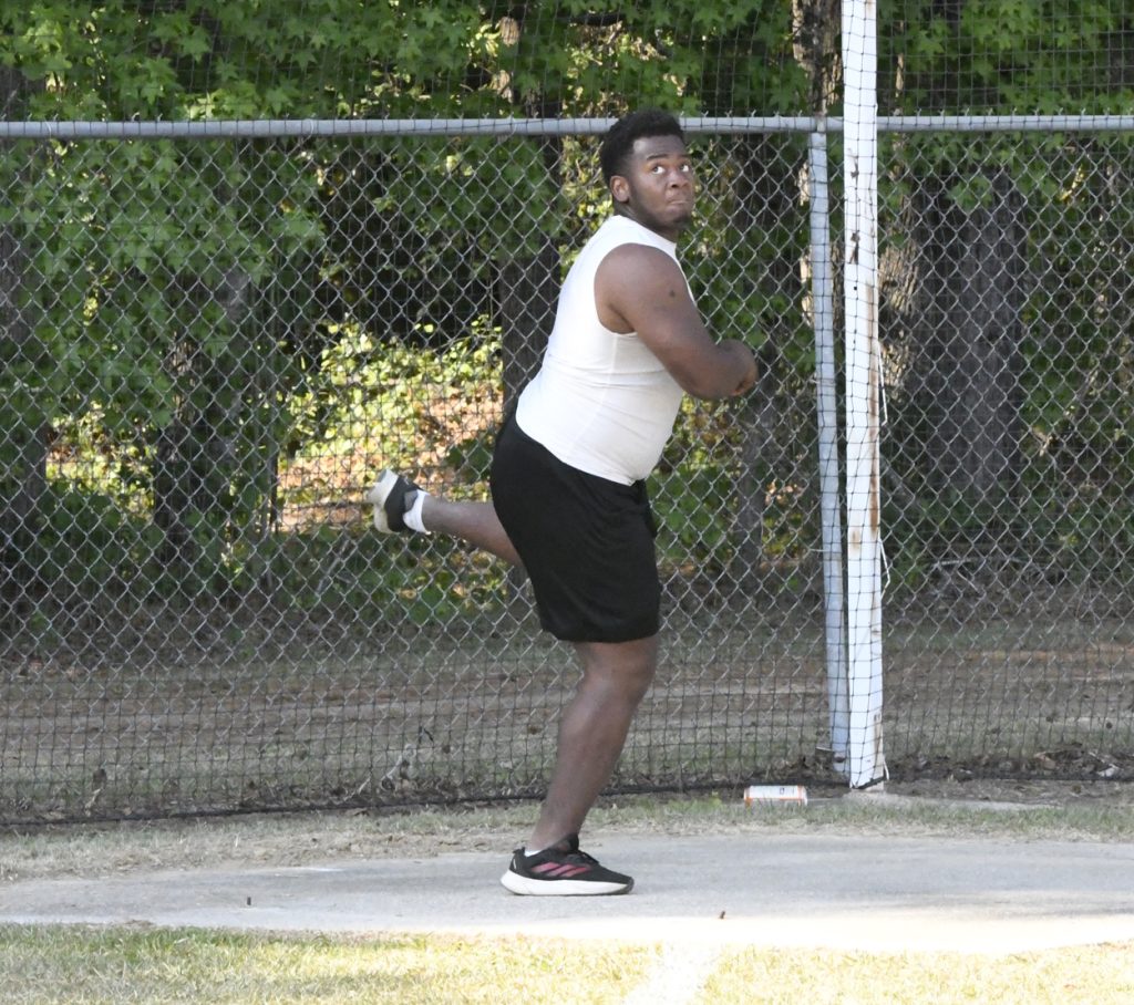 A person in athletic wear throws a discus on an outdoor track field, fenced area and green trees in the background.