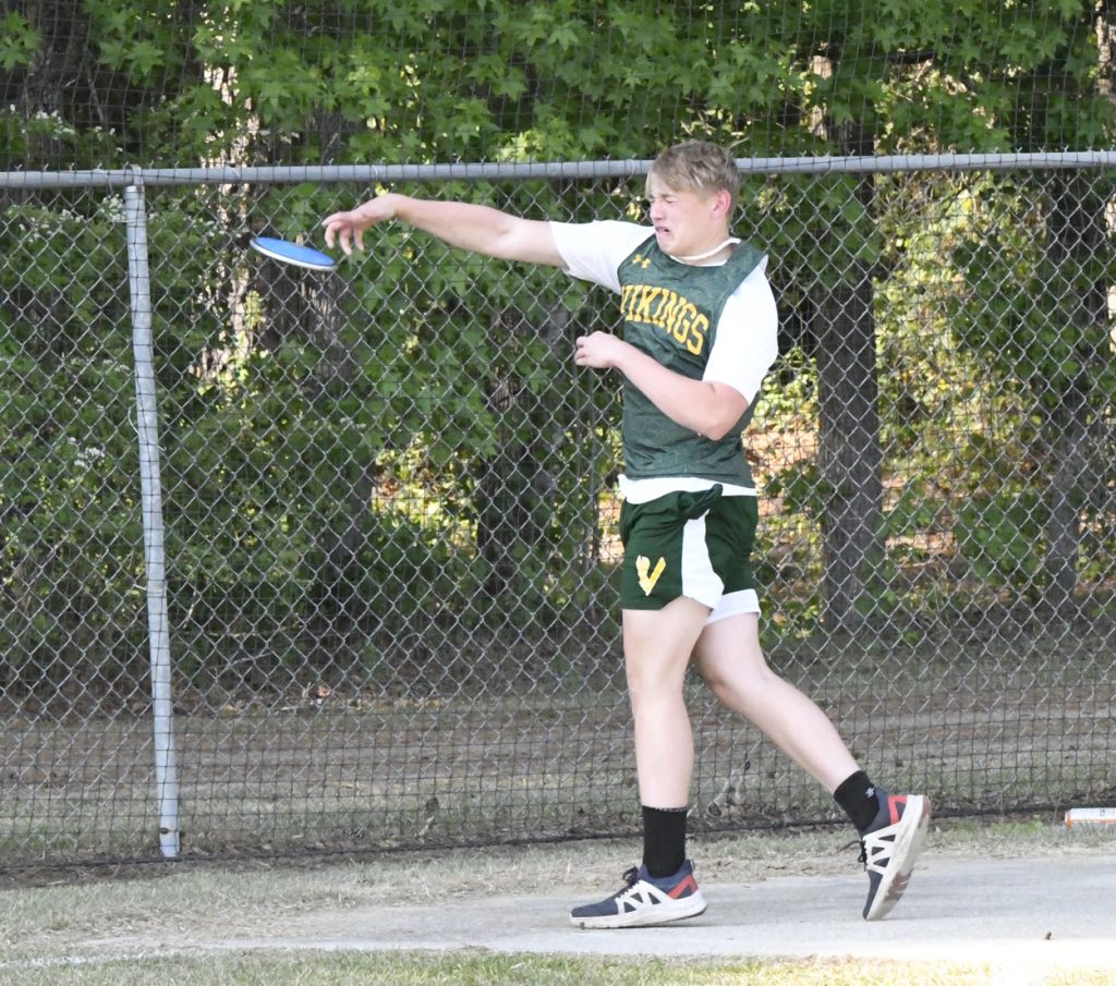 A young athlete in a green "Vikings" uniform throws a discus during an outdoor track and field event, with a chain-link fence and trees in the background.