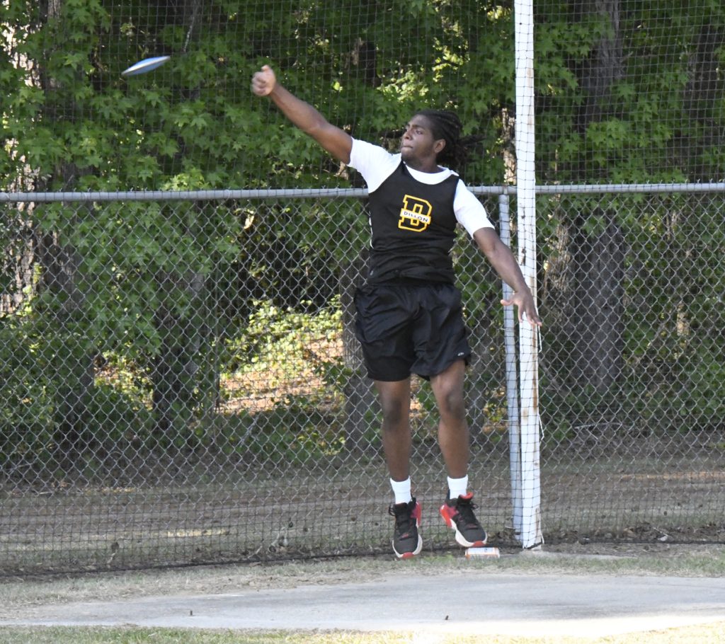 A male athlete in a black uniform throws a discus during an outdoor track and field event by a chain-link fence and trees.