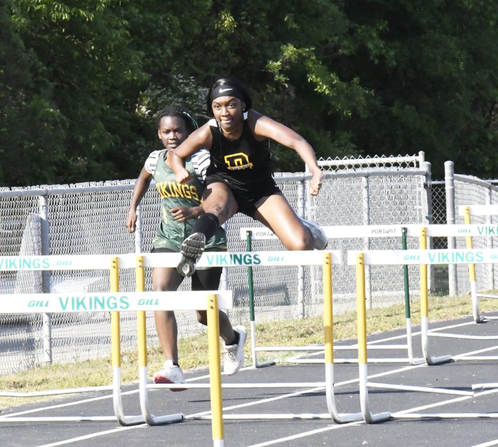 Two female athletes race in a hurdles event on an outdoor track, with one athlete in the lead, both clearing hurdles.