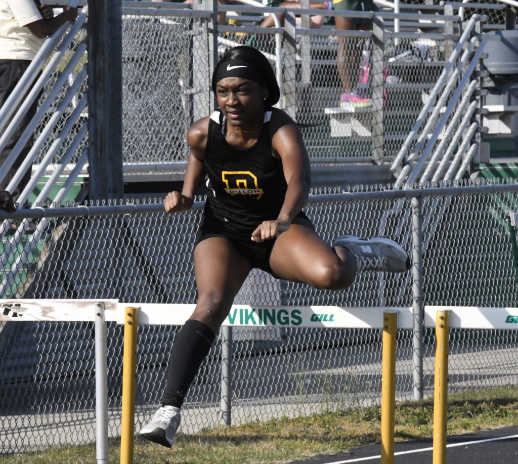A female athlete in black sportswear jumps over a hurdle during a track and field race, with stadium bleachers in the background.