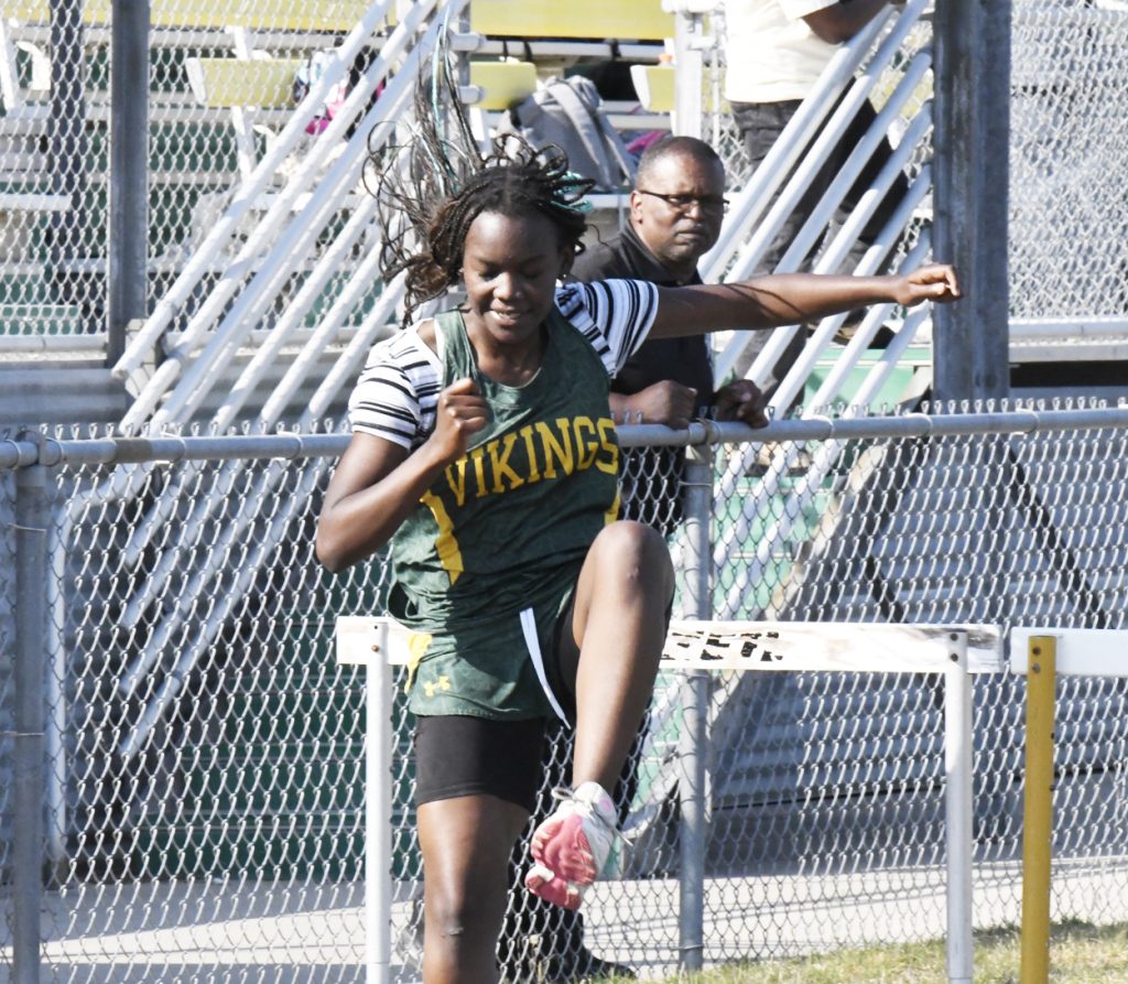 A female athlete wearing a green "Vikings" jersey jumps over a hurdle during a track and field event.