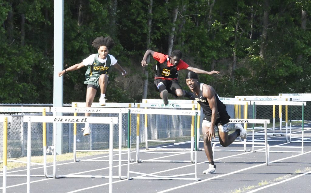 Three athletes compete in a hurdle race on a track, each mid-jump over hurdles, with trees and fencing in the background.