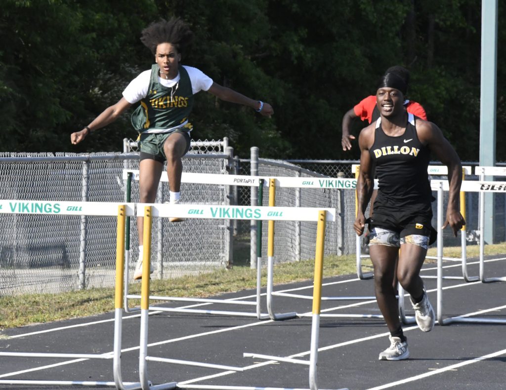 Two male athletes compete in a hurdles race on an outdoor track, one jumping over a hurdle and the other running past it.
