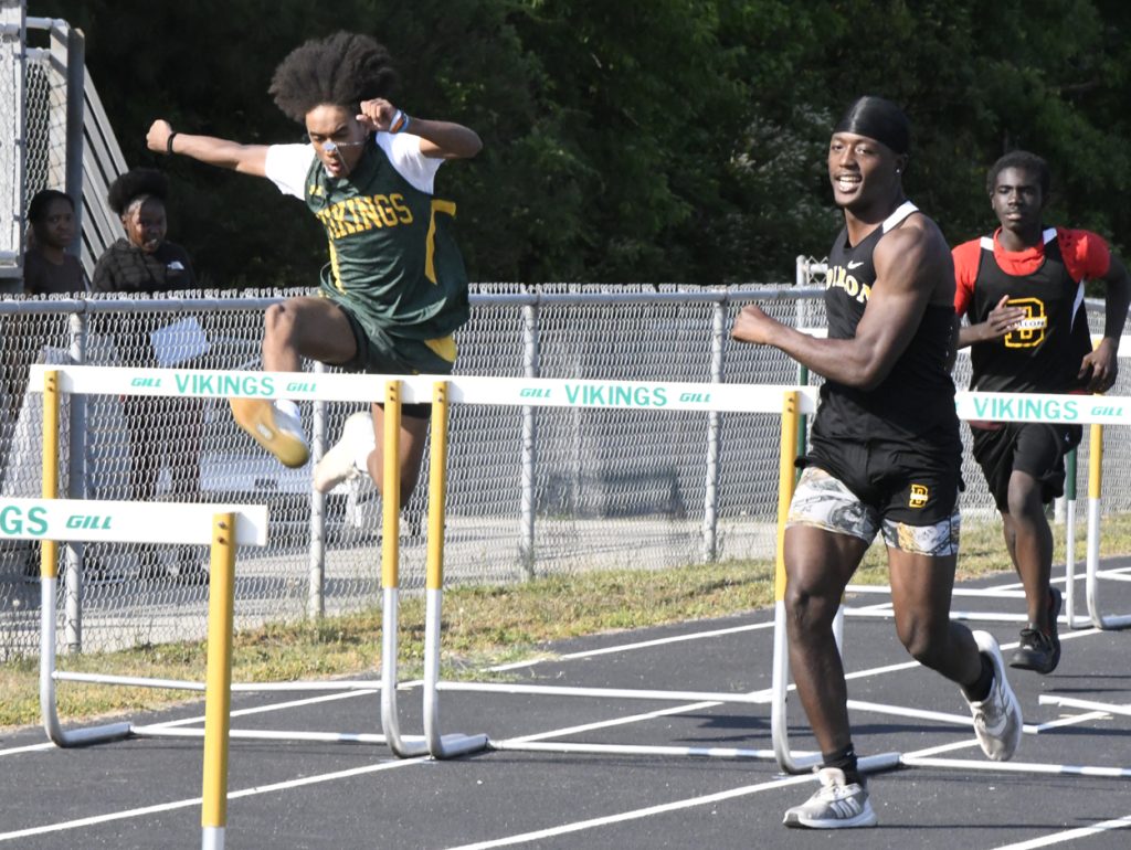 Athletes compete in a hurdles race on an outdoor track, with one runner mid-jump and others sprinting, spectators watch from behind a chain-link fence.