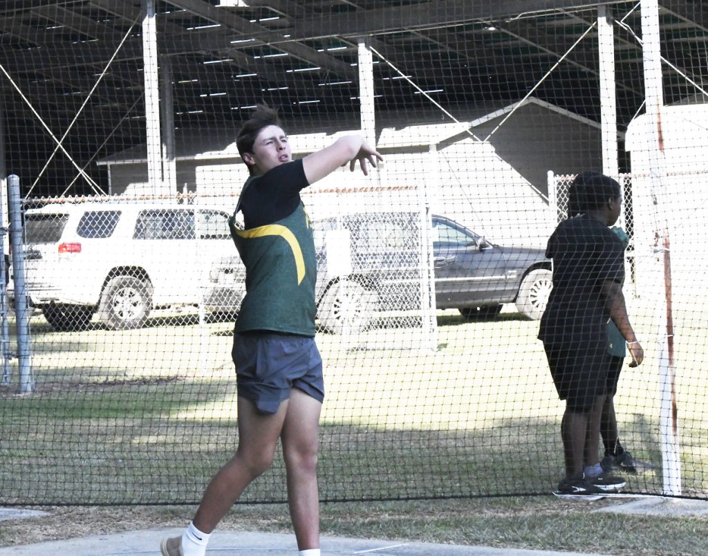 A young athlete in a green and yellow sports uniform throws a discus inside a fenced area, while two others stand nearby. Vehicles are parked in the background.