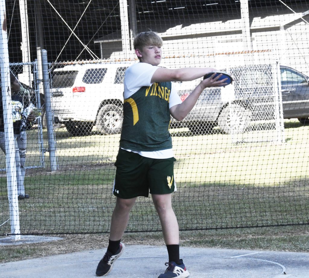 A young athlete wearing a green "Vikings" uniform prepares to throw a discus during a track and field event, standing inside a netted throwing area.