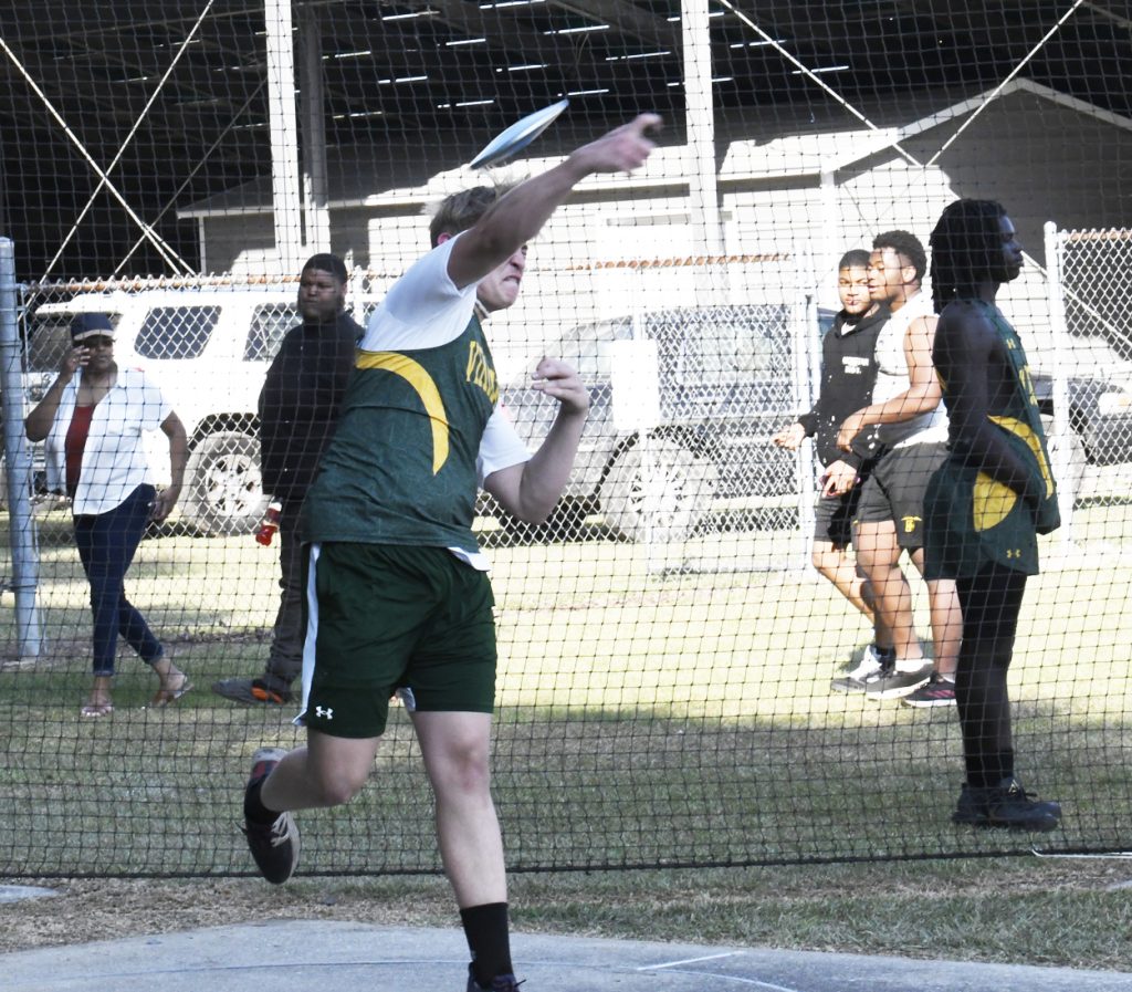 A male athlete in green sportswear throws a discus during a track and field event, with spectators and other athletes watching behind a net in the background.