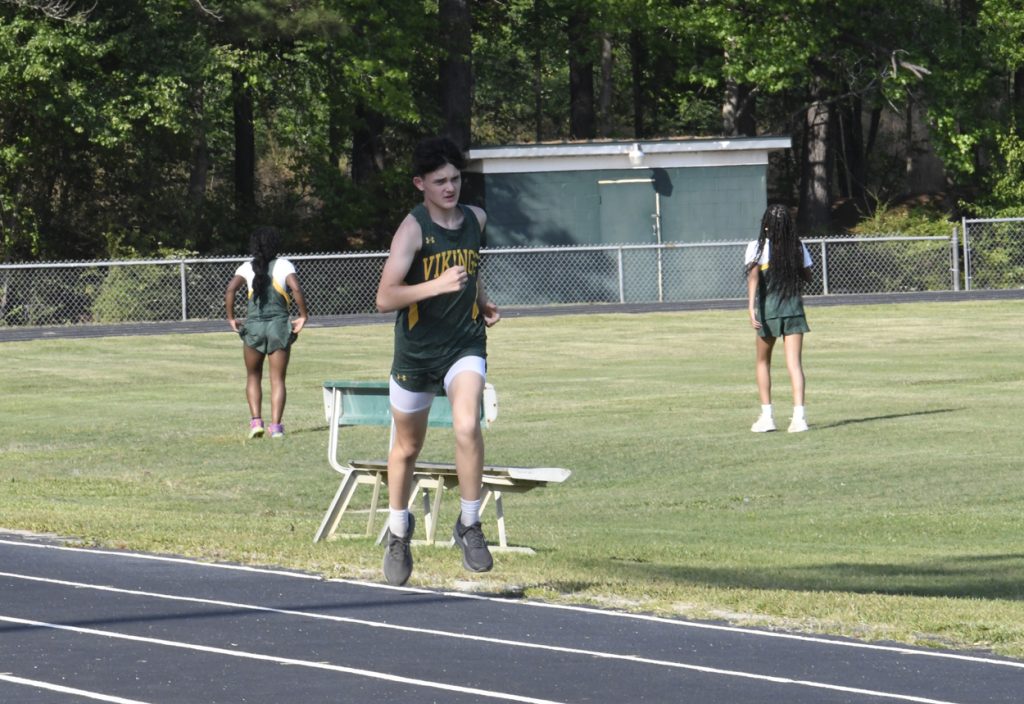 A student in a green Viking uniform runs on a track, with two other students and a bench on the grassy field behind him.